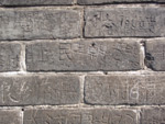 Valokuva harmaasta kivimuurista, jonka kivien välissä valkoista laastia. Kivimuurin kivet ovat symmetrisiä ja tiilenmuotoisia. Kiviin kaiverrettu kiinalaisia kirjaimia.