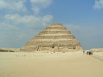 Aavikolla seisova vanha pyramidi, joka on pilvistä sinitaivasta vasten elottomalla vaalealla aavikolla. Pyramidi on monikerroksinen ja rapistunut, maalaamaton suuri kolmionmuotoinen rakennus.