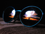Valokuva aurinkolaseista tumman karkean kankaan päällä. Aurinkolasien peililinsseistä heijastuu iltarusko ja sen keskellä suuri valkoinen sienipilvi. Ydinräjähdyksen sienenmuotoista pilveä muistuttava pilvi heijastuu molemmista linsseistä.