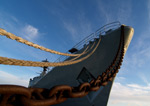 Valokuva sotalaivasta, fregatista, joka on ankkuroitu köysin ja ketjulla satamaan. Fregatti on kuvattu alhaalta viistosti ylöspäin, joten alus näyttää suurelta sinistä pilvitaivasta vasten.