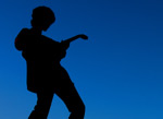 Valokuva kitaristin mustasta siluetista hyvin voimakkaasti tummansiniseksi värjättyä hämärää taustaa vasten. Kitaristi on vasemmalla soitellen kummallakin kädellä instrumenttiaan.