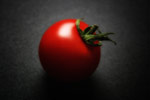 Punainen tomaatti, jossa vihreä kuivuneista lehdistä muodostunut kanta. Tomaatti on kirkkaanpunainen ja se nököttää yksin tummanharmaata taustaa vasten. Kuvan laidat ovat tummemmat kuin keskiosa.