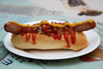 Hotdog valkoisella paperilautasella. Hotdogissa on suuri makkara, jonka välissä kellertävä leipämäinen osa. Hotdogin päällä on punaista ketsuppia, sinappia ja sipulia sun muita hotdog-mausteita.