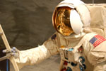 Valkoiseen avaruuspukuun pukeutunut astronautti pitää toisella kädellään kiinni jostakin valkoisesta metalliputkesta. Astronautilla kullanvärinen naamio kasvojensa edessä ja astronautilla myös olkapäässä Yhdysvaltain lippu.