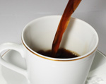 Valkoinen kahvikuppi melkein puoliksi täynnä mustaa kahvia, jota valuu norona kupin keskelle. Kahvi on melkein mustaa eikä sen seassa ole maitoa tms.