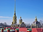 Kuva Pieterista. Kuvassa sininen lähes täysin pilvetön taivas ja Pietarin suuria taivaita kurkottavia torninhuippuja. Kuvan alaosasa punaiseksi maalattuja rakennusten seiniä Pietarissa.