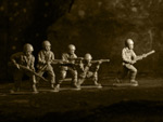 Lauma sotaukkoja rivissä juoksemassa vasemmalta oikealla ruskeasävyisessä valokuvassa. Kaikki sotaukkelit ovat erinäköisiä, sillä osa juoksee ja toiset ovat polvillaan tai seisovat ase kädessä tähdäten oikeaan suuntaan.